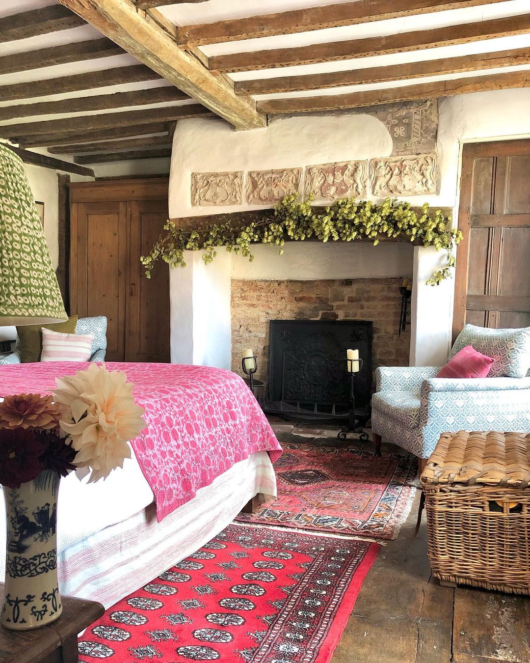 Historic bedroom with exposed wooden beams, fireplace with hop garland decoration, pink bedding, patterned rugs, and traditional furnishings