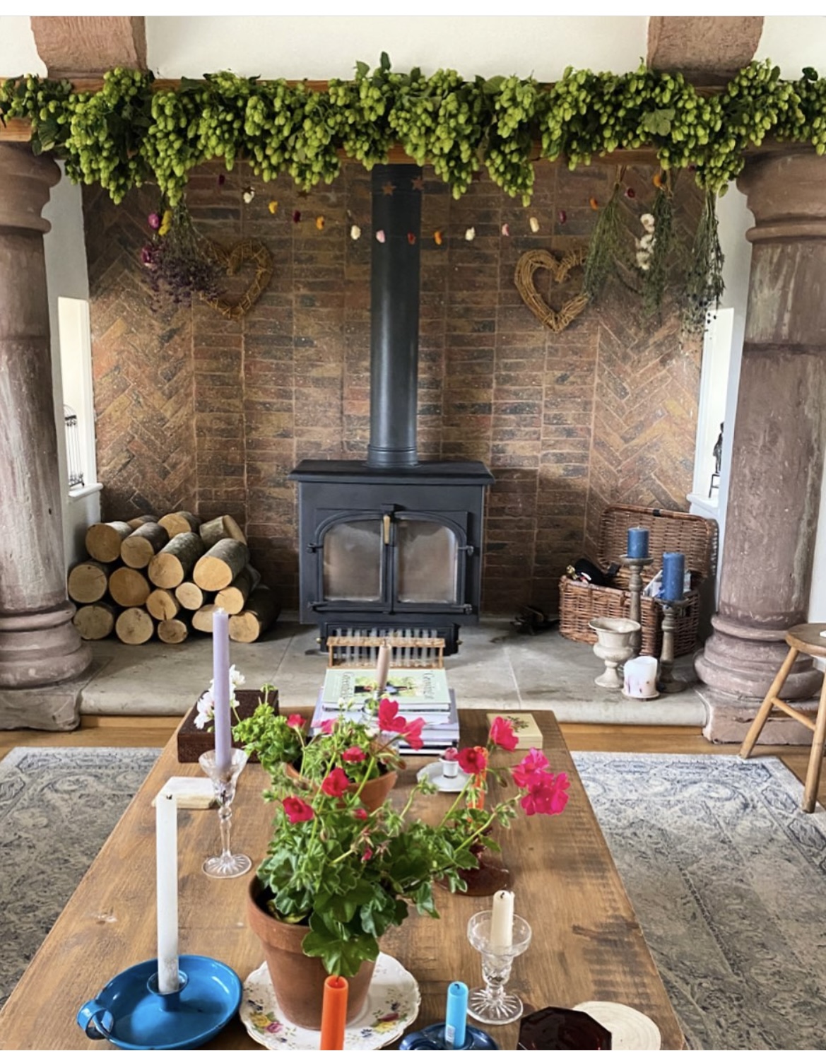 Fireplace with black wood stove, hop garland mantel decoration, wooden table with pink flowers, candles, and brick herringbone wall