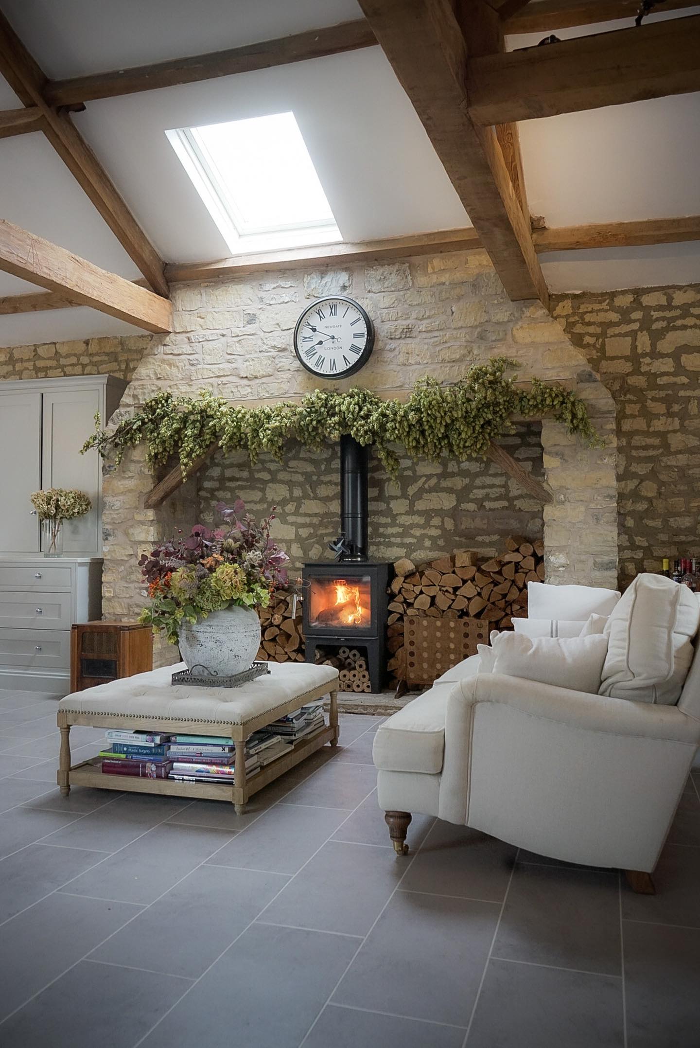 Modern farmhouse living room with exposed wooden beams, stone fireplace, black wood stove, cream armchairs, hop garland mantel decoration, and skylight