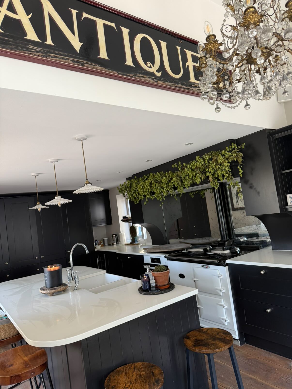 Modern kitchen with black cabinetry, white countertops, island with wood stools, pendant lights, crystal chandelier, and hop garland decoration above black wall