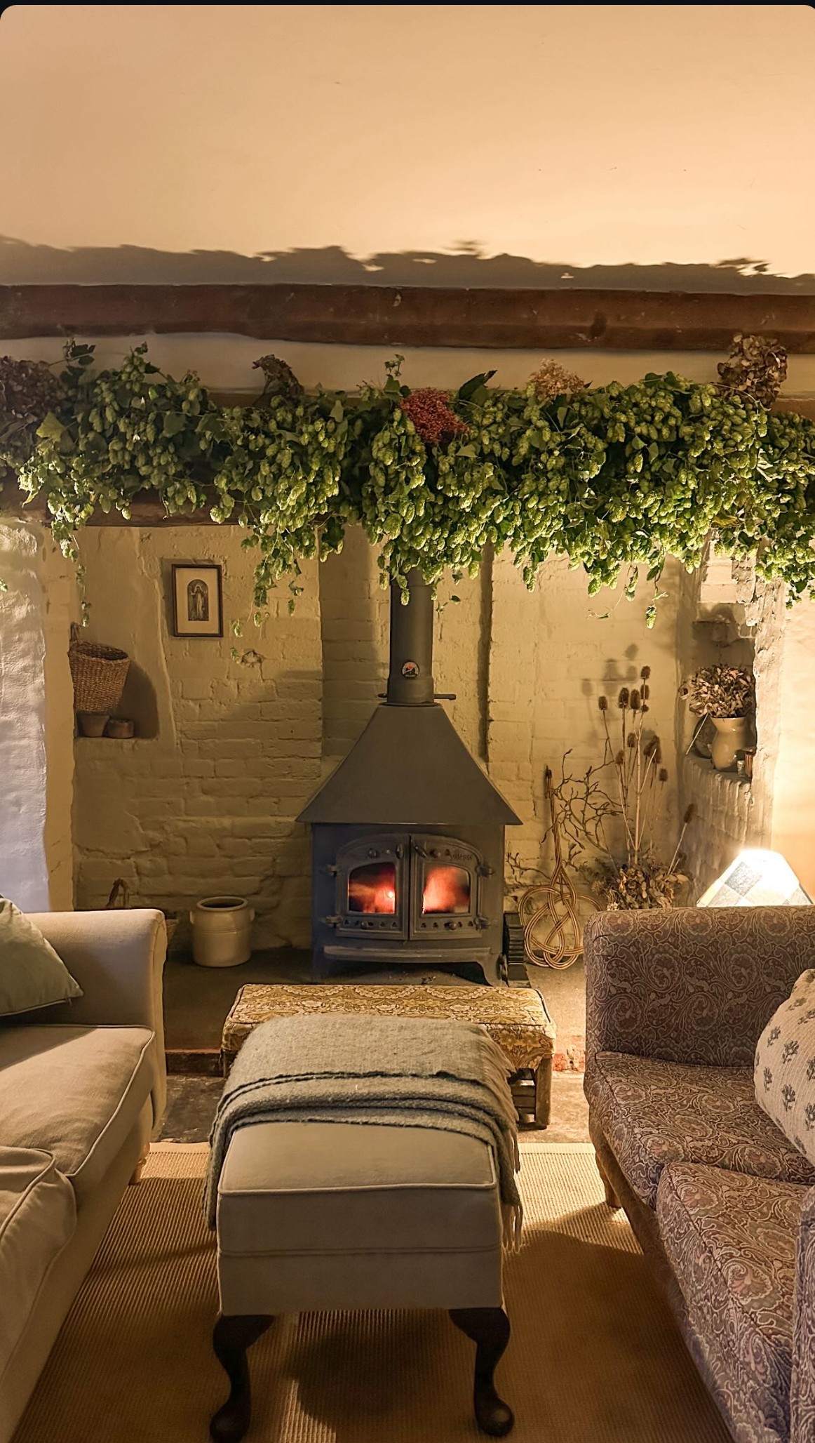 Cozy living room with wood-burning stove, comfortable seating, hop garland decoration above fireplace, and view of water and mountains beyond