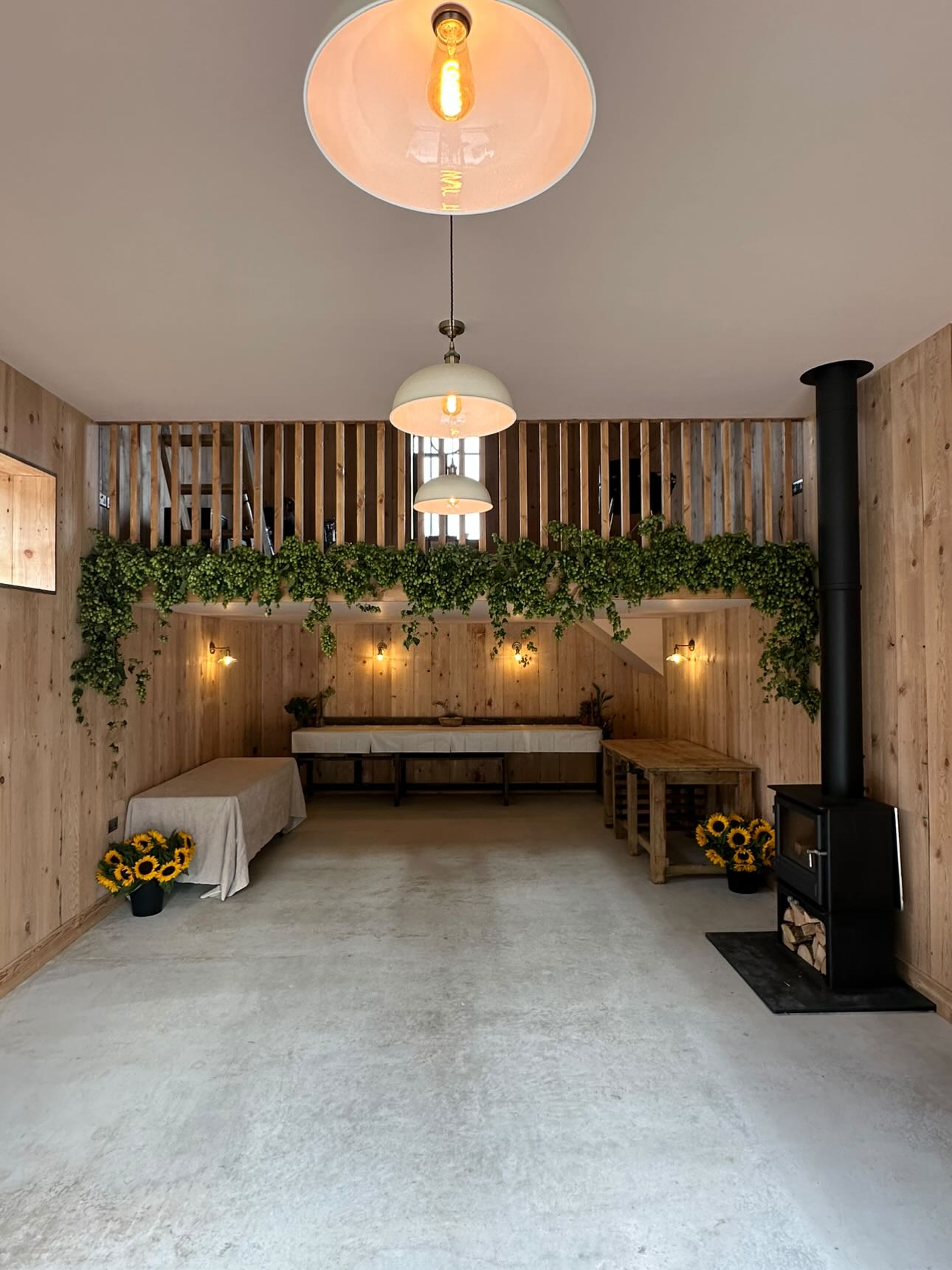 Modern barn interior with wooden walls, concrete floor, pendant lights, hop garlands, sunflowers, fireplace, and wooden furniture for events