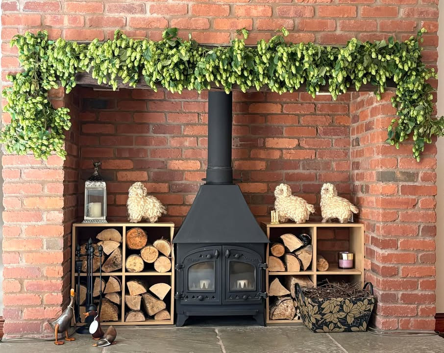Black wood stove with firewood storage cubbies, decorative sheep figurines, and hop garlands draped above red brick fireplace