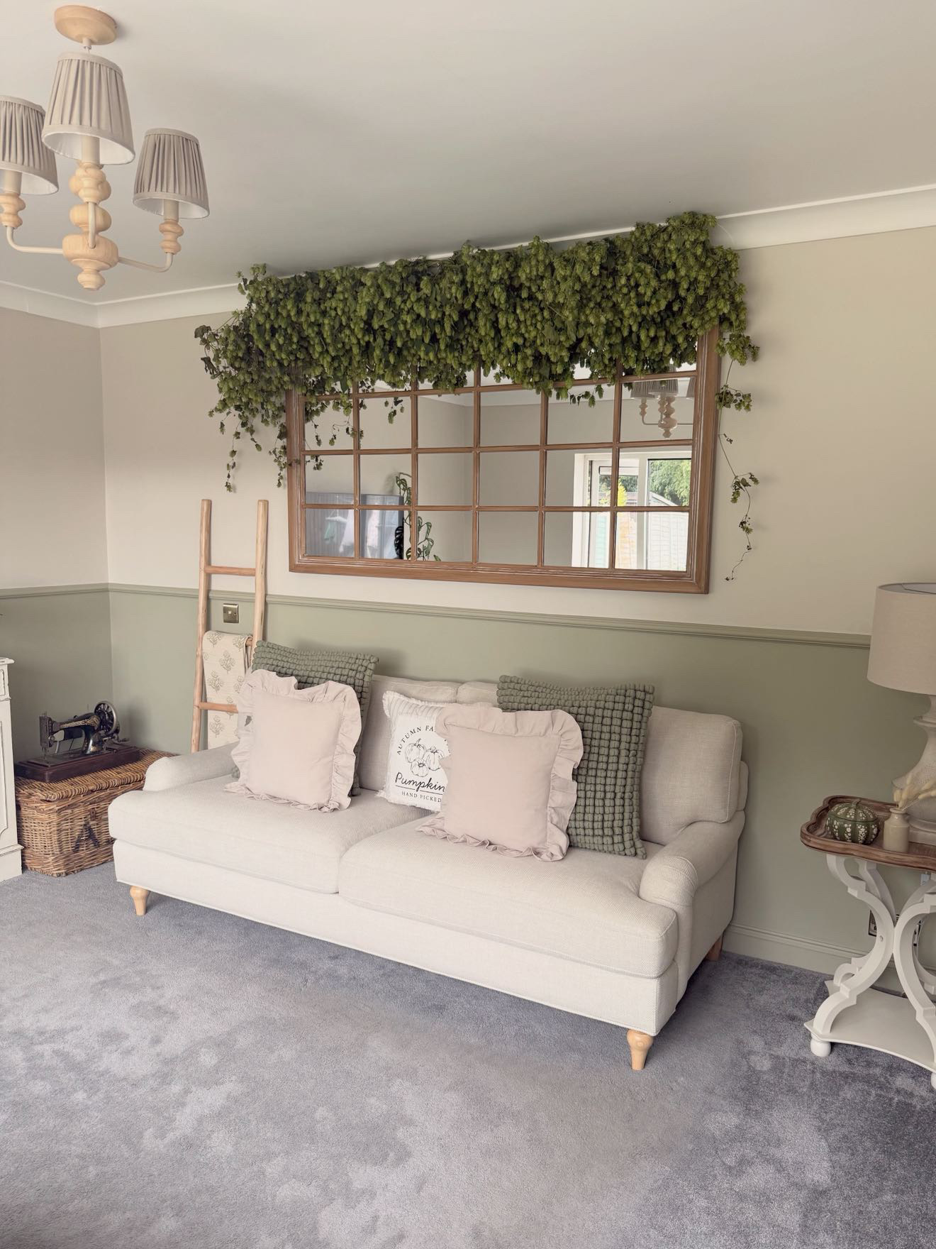 Cozy living room with cream sofa, wooden window frame with cascading hop garland, wooden ladder, wicker basket, pendant lights, and two-tone sage green and crea