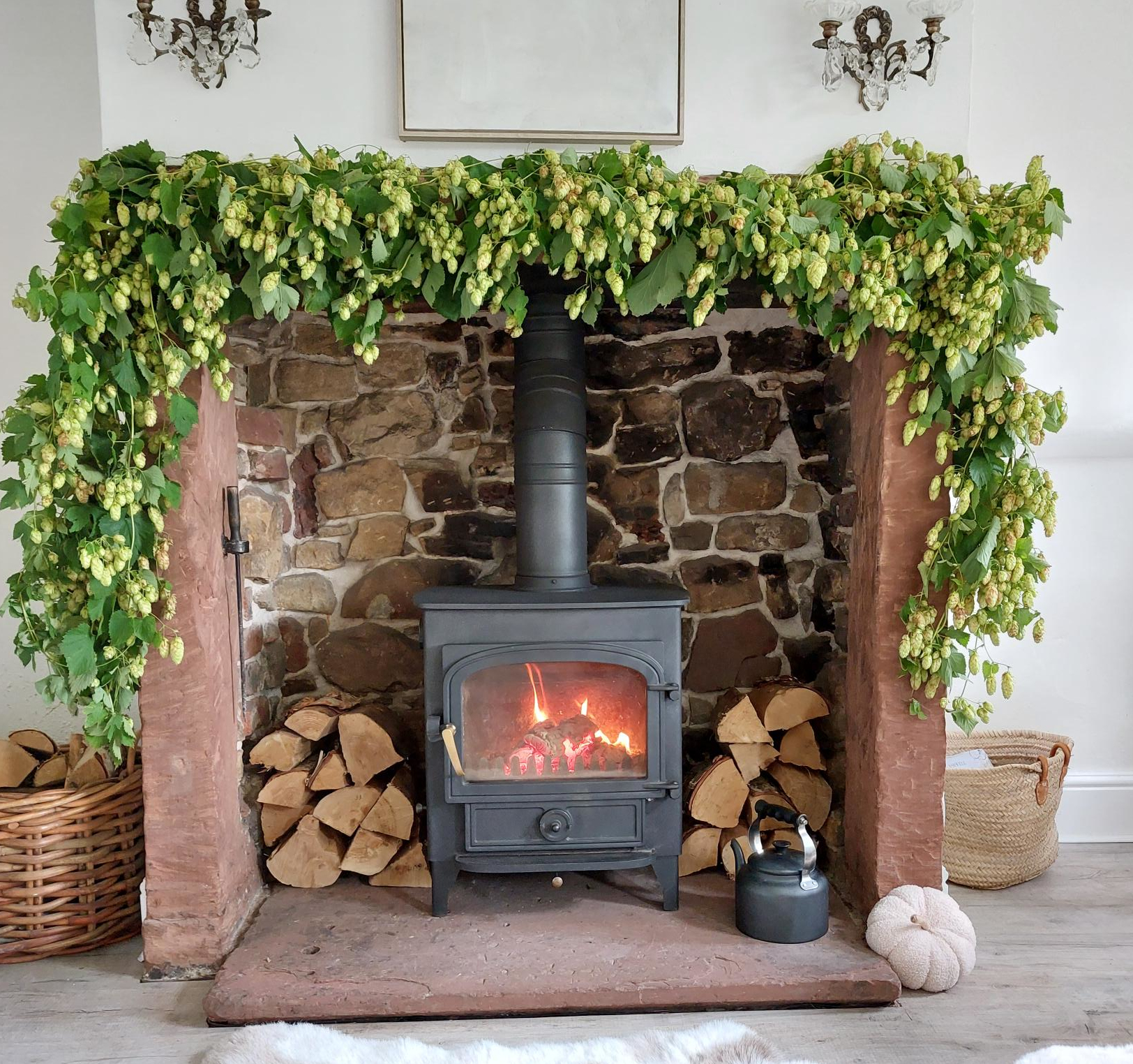 Black wood-burning stove with glowing fire in stone fireplace decorated with hop garlands, stacked firewood, wicker baskets, and decorative accessories