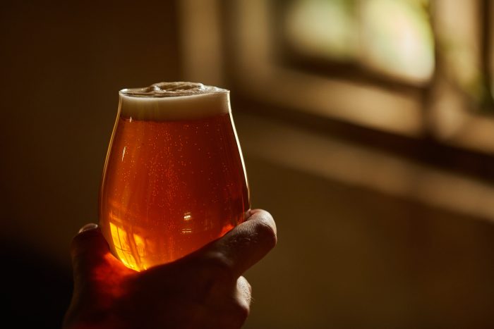 Hand holding a glass of amber beer with foam head, backlit by warm sunlight casting a golden glow through the liquid